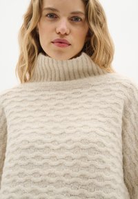 Beige Pullover mit hohem Kragen und strukturiertem Wellenmuster, aus weichem, flauschigem Material gefertigt. Mit dicker gerippter Kragen. Minimalistisches Design.