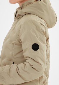 Beige puffjacka med huva, med en kviltad textur och en svart cirkulär logotyp patch på vänster ärm. Inkluderar framfickor med dragkedja.