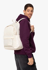 Sac à dos blanc au design arrondi, texture en tissu, avec des doubles glissières et une poche avant, porté sur un sweat à capuche violet par une personne.
