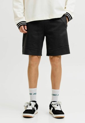Persoon draagt zwarte knielange shorts, witte hoge sokken met groene tekst, zwart-witte sneakers en een witte top met lange mouwen met zwart gestreepte boordjes.