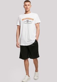 Hvit bomull T-skjorte med "Gryffindor" tekst i oransje og svart, sammen med svarte mesh-shorts og hvite joggesko.