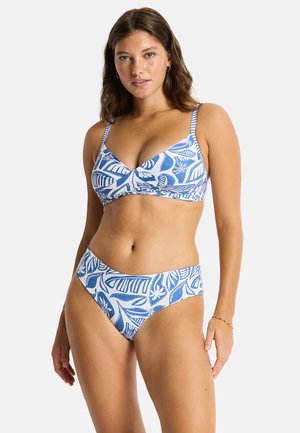 Kobieta w niebiesko-białym bikini w liściasty wzór z regulowanymi ramiączkami, stojąca na jasnym jednolitym tle.