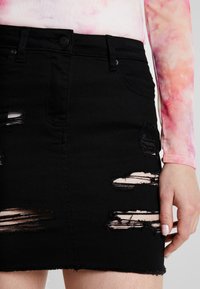 Jupe en denim noir avec plusieurs zones usées, une coupe ajustée, un ourlet brut et des poches avant. Le tissu a une texture lisse.