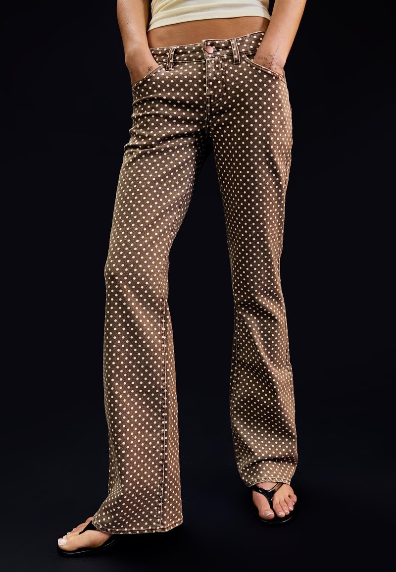 Femme portant un jean flare marron à pois blancs, les mains dans les poches, un haut beige et des sandales noires à semelles plates, sur fond noir.