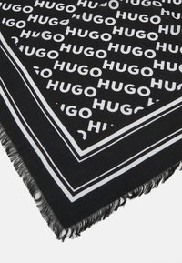 Svart skjerf med hvit "HUGO"-tekst som gjentas. Har et dristig design med frynser langs kantene og hvite kanter som rammer inn mønsteret.