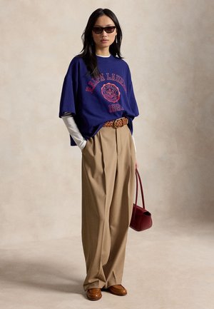 Frau trägt ein dunkelblaues Ralph Lauren T-Shirt über weißen Langarmshirts, beige weit geschnittene Hosen mit geflochtenem Gürtel, braune Schuhe, Sonnenbrille und hält eine rote Handtasche.