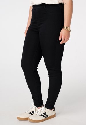 Femme portant un jean skinny noir, des baskets blanches avec des rayures noires, un haut blanc et des bracelets dorés, debout devant un fond clair uni.
