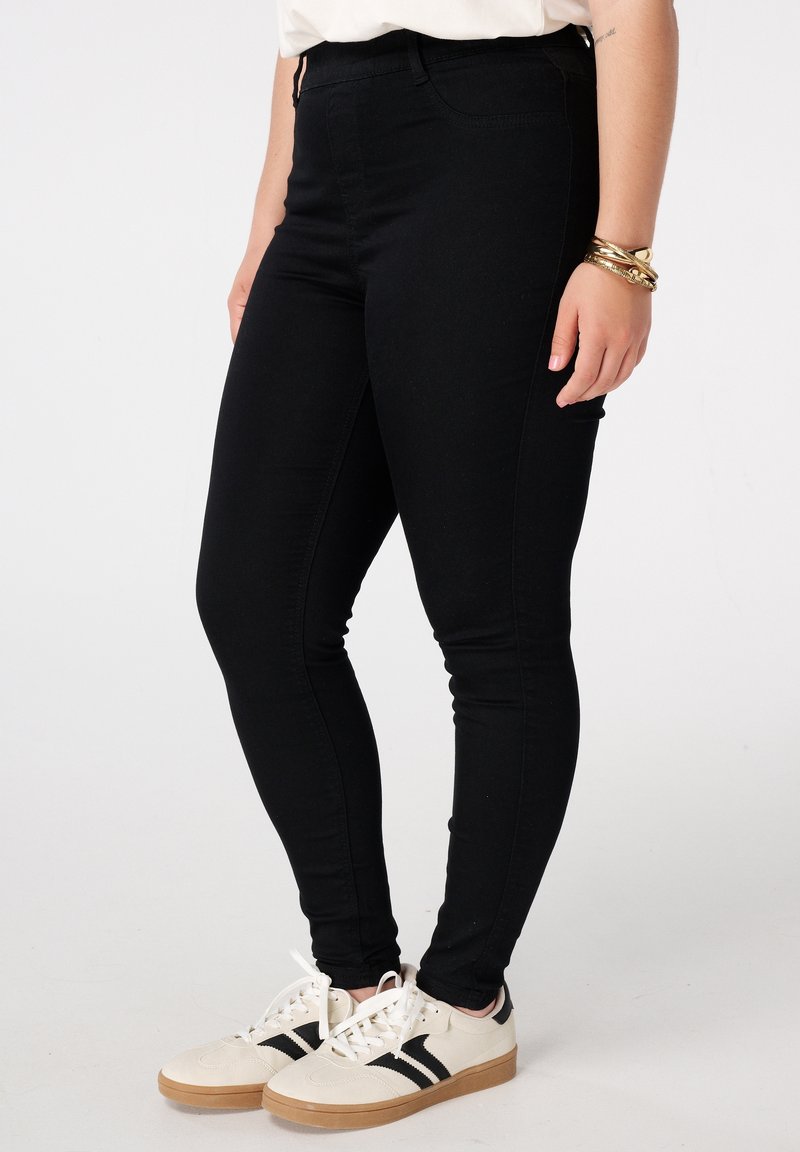 Femme portant un jean skinny noir, des baskets blanches avec des rayures noires, un haut blanc et des bracelets dorés, debout devant un fond clair uni.