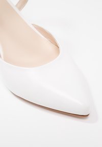 Chaussure à talon haut blanche à bout pointu avec dessus en cuir lisse et doublure intérieure nude sur fond blanc.