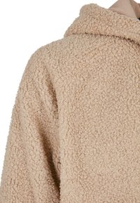 Beige Kapuzenpullover aus weichem, flauschigem Material. Er verfügt über einen lockeren Schnitt mit einem dezenten Muster und minimalen Nähten.
