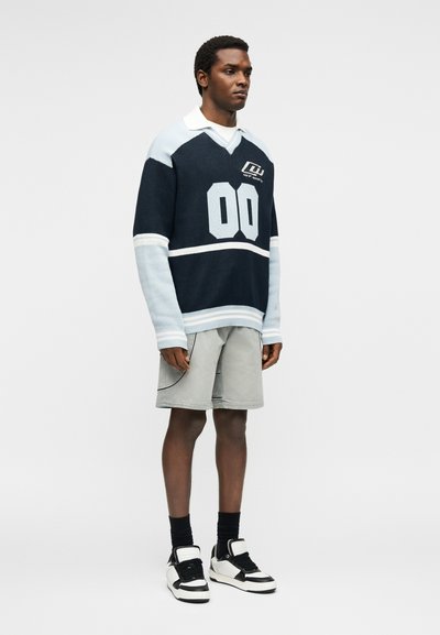 Άνδρας που φοράει μπλε σκούρο και ανοιχτό oversized πουλόβερ με τον αριθμό 00, ανοιχτό γκρι σορτς, μαύρες κάλτσες και ασπρόμαυρα αθλητικά, στέκεται ακίνητος.