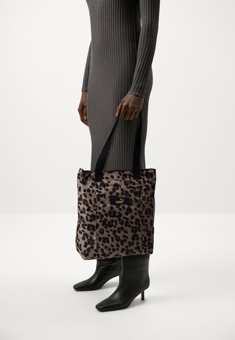 Personne portant une robe grise côtelée et des bottes à talons noires tient un sac fourre-tout Puma imprimé léopard avec des poignées noires.