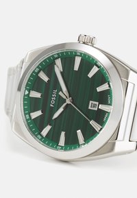 Montre en acier inoxydable argenté avec un cadran à rayures vertes, des index horaires blancs et un affichage de la date. Elle possède un bracelet en métal brossé et une couronne.