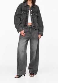 Chaqueta de mezclilla negra y jeans de pierna ancha con un acabado desgastado. Presenta grandes bolsillos y una superficie texturizada. Se lleva con sandalias negras de tacón.