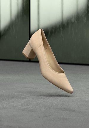 Zapato pump de tacón bloque de ante beige con punta puntiaguda, mostrado flotando sobre una superficie gris frente a un fondo desenfocado verde y negro.