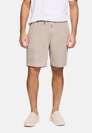 Hombre con pantalones cortos de lino beige con cintura ajustable y zapatos beige sin cordones, de pie frente a un fondo liso.