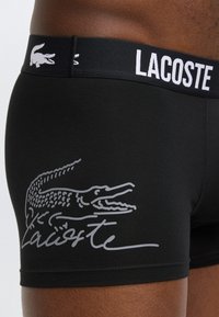 Μαύρα ανδρικά εσώρουχα Lacoste με λευκό κείμενο λογότυπου στη μέση και μεγάλο γραφικό κροκόδειλο στο αριστερό ισχίο, φορεμένα από άτομο με σκούρο δέρμα.