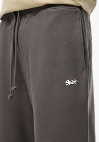 Mørkegrå sweatpants med en blød tekstur, der har et hvidt logo på det nederste ben og en snor i taljen for justerbar pasform.