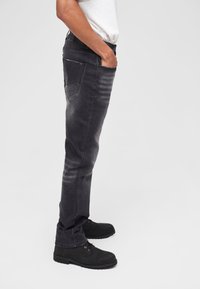 Mustad teksased musta värvi, kulunud viimistlusega, sirge lõike ja viie taskuga stiiliga, kus õmblused on selgelt nähtavad. Sobitatud mustade paeltega saapadega.