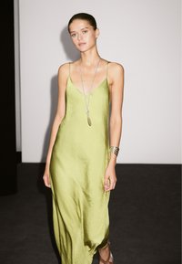 Satin-Spaghetti-Top-Kleid in Hellgrün, mit glatter Textur und subtiler glänzender Oberfläche. Accessoires sind eine lange silberne Halskette und gestapelte Armreifen.