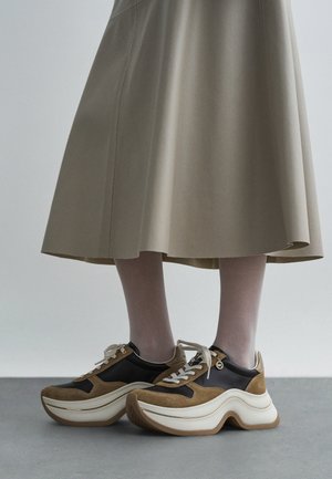 Sko med en tykk, lagdelt såle i beige og brunt semsket skinn, med en svart læroverdel, hvite lisser og en gullfarget detalj.