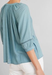 Blouse bleu clair à manches trois-quarts, tissu texturé, col froncé et détails en dentelle aux ouvertures des manches. Portée avec un pantalon blanc.