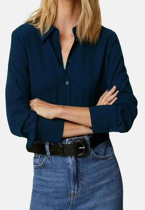 Vrouw met gekruiste armen, gekleed in een donkerblauw overhemd met gestructureerde mouwen en een hoge taille blauwe spijkerbroek met een zwarte riem.
