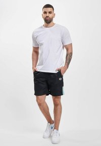 Pantaloni scurți de baie negri cu un accent turcoaz, asortați cu un tricou alb simplu. Modelul stă în adidași albi, expunând un tatuaj pe braț.