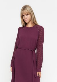 Rochie cu mâneci lungi într-o nuanță profundă de mov, cu un material texturat. Prezintă o talie ajustată și un cordon asortat, cu mâneci sheer cu un model.