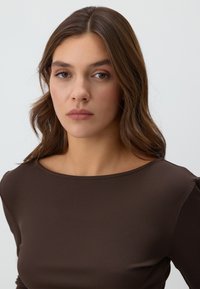 Bruine geribbelde langarmtop met een wijde halslijn en een soepele textuur, met natuurlijk haar dat het gezicht omlijst tegen een lichte achtergrond.