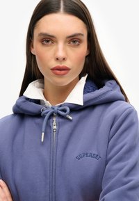 Sudadera morada con cremallera y capucha ajustable, textura suave, logo bordado y una camisa blanca de cuello debajo.