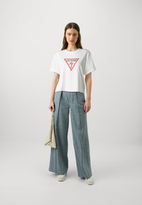 T-shirt en coton blanc avec un logo triangulaire rouge portant l'inscription "GUESS?", associé à un pantalon en denim à jambes larges et un sac à bandoulière beige clair.