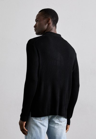 Svart ribbad tröja med långa ärmar, har en vikbar krage och en avslappnad passform. Bärs med ljusblå denimjeans.