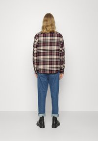 Camicia di flanella a scacchi nei colori beige, bordeaux e nero, con maniche lunghe e orlo arcuato. Abbinata a pantaloni in denim blu con risvolti e stivali neri.
