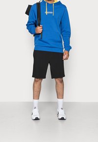 Blå hoodies med gula accenter, svarta shorts, vita sneakers och vita strumpor. Har en logotyp på bröstet och en framficka.