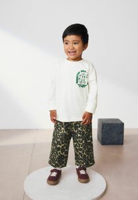 Chemise blanche à manches longues avec un imprimé graphique vert, associée à un pantalon ample imprimé léopard vert olive et des baskets bordeaux avec des lacets blancs.