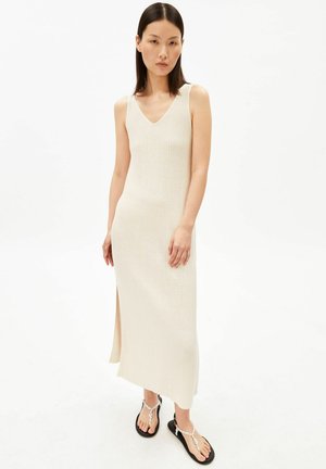 Femme aux cheveux lisses et foncés portant une robe midi beige côtelée sans manches avec une fente latérale et des sandales noires à brides blanches.