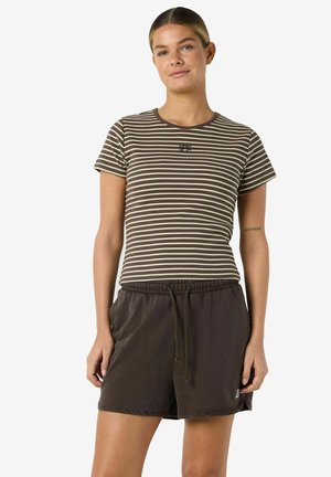 Nő barna és bézs csíkos rövid ujjú ingben és barna húzózsinóros rövidnadrágban, kezei lazán oldalán áll.