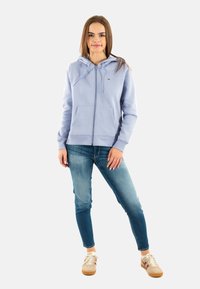 Felpa azzurra chiaro con cerniera, cappuccio e tasche frontali, abbinata a jeans blu aderenti e sneakers beige. Tessuto liscio, design casual.