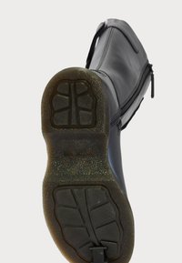 Bota negra con suela de goma texturizada vista desde la parte inferior, resaltando las bandas de agarre y la cremallera lateral sobre un fondo claro.