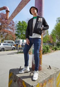 Casaco bomber preto com a letra "R", camiseta branca de gola redonda, jeans skinny azuis e ténis brancos de cano alto; de pé sobre um bloco de concreto.