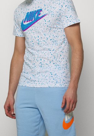 T-shirt Nike blanc à taches bleues avec logo bleu, associé à un short bleu clair avec un swoosh Nike orange.