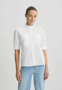Chemise blanche à manches courtes avec col mandarin, dotée d'une fermeture à boutons et de détails en panneaux sur le devant supérieur. Portée avec un jean bleu clair.