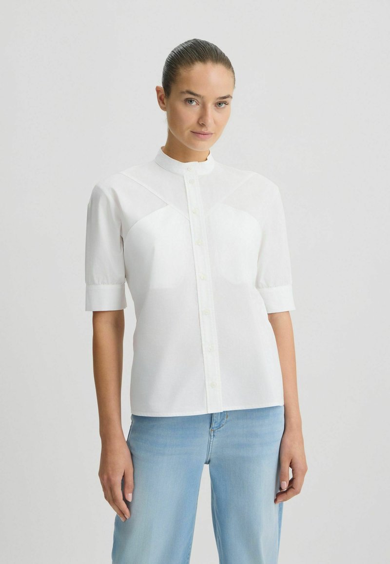 Chemise blanche à manches courtes avec col mandarin, dotée d'une fermeture à boutons et de détails en panneaux sur le devant supérieur. Portée avec un jean bleu clair.