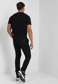 Hombre con camiseta negra, pantalones negros con bolsillos traseros y zapatillas en blanco y negro, de pie contra una pared blanca en la esquina.