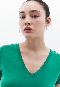 Camiseta verde de cuello en V en tela suave, con forma ajustada y mangas cortas. Primer plano que muestra el tono de piel y los pendientes, contra un fondo blanco.