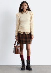 Light beige sweater with puffed sleeves layered over a beige and brown plaid shirt. Brown plaid skirt, burgundy socks, and black chunky boots. Brown handbag.