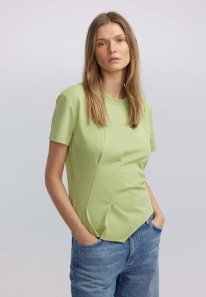 Camiseta de manga corta verde claro con un diseño ajustado y acentos texturizados, combinada con jeans azules claros. Cuello simple, estilo casual.