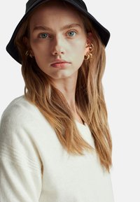 Frau trägt einen cremefarbenen strukturierten Pullover und einen schwarzen Bucket Hat. Goldene Creolen und lange glatte Haare sind sichtbar.