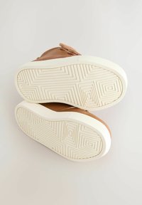 Hnědé kožené boty s páskem na suchý zip, vybavené texturovanou bílou gumovou podrážkou s geometrickými vzory. Design je nízký a neformální.
